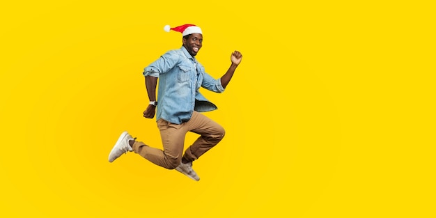 Portret van de volledige lengte van een gelukkige vrolijke man met een kerstmuts van het nieuwe jaar die springt of vliegt, haast je naar zijn droom, kijkend naar de camera met een brede glimlach. indoor studio-opname geïsoleerd op gele achtergrond