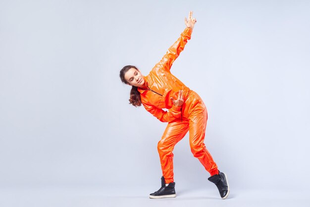 Portret van de volledige lengte van een positief energiek tienermeisje met donkerbruin haar in een feloranje jumpsuit die hiphop danst en omhoog wijst met een vingerpistool, hobbyactiviteiten. studio opname, witte achtergrond