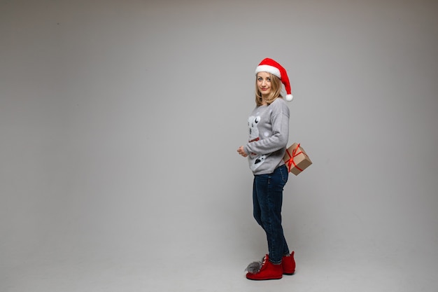 Foto portret van de volledige lengte van mooie blonde vrouw in rode kerstmuts, trui, jeans en laarzen met cadeau achter haar rug en duim opdagen ter goedkeuring