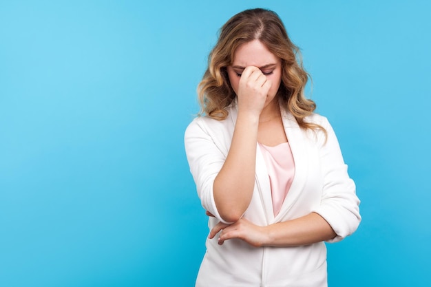 Portret van depressieve dramatische vrouw met golvend haar in witte jas huilend in ogen wrijvend, wanhopig en hopeloos voelend, bezorgd over verlies, hoofdpijn lijdend. indoor studio-opname, blauwe achtergrond