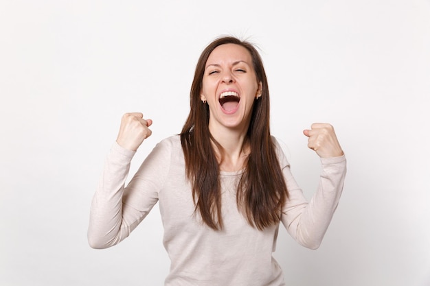 Portret van dolblij schreeuwende jonge vrouw in lichte kleding ogen gesloten houden winnaar gebaar geïsoleerd op een witte muur achtergrond. Mensen oprechte emoties, lifestyle concept. Bespotten kopie ruimte.