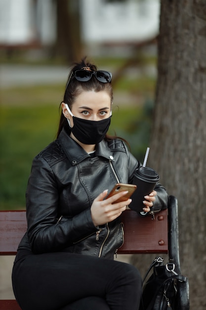 Portret van donkerbruine vrouw in een zwarte beschermende maskerzitting op een bank in het park, zonnige de lentedag. Vrouw praten op mobiele telefoon