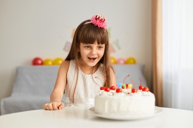 Portret van donkerharige opgewonden gelukkig klein meisje kijkend naar verjaardagstaart viert haar vakantie uiting van positieve emoties poseren met feestelijke inscriptie en ballonnen op achtergrond