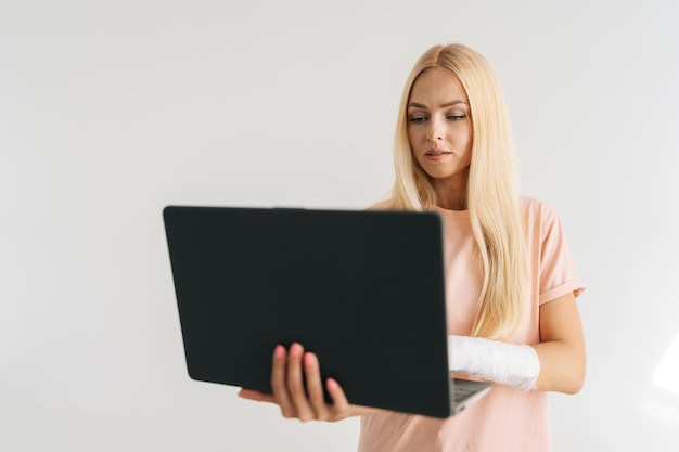 Portret van doordachte gewonde jonge vrouw met gebroken arm gewikkeld in gipsverband met laptop en werkend typen staande op een witte geïsoleerde achtergrond