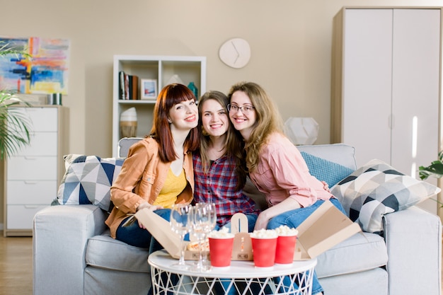 Portret van drie gelukkige jonge vrouwelijke vrienden die huispartij met pizza, popcorn en wijn hebben, die op de grijze bank in eetkamer thuis zitten. Pizzaparty, vrouwelijke vriendschap