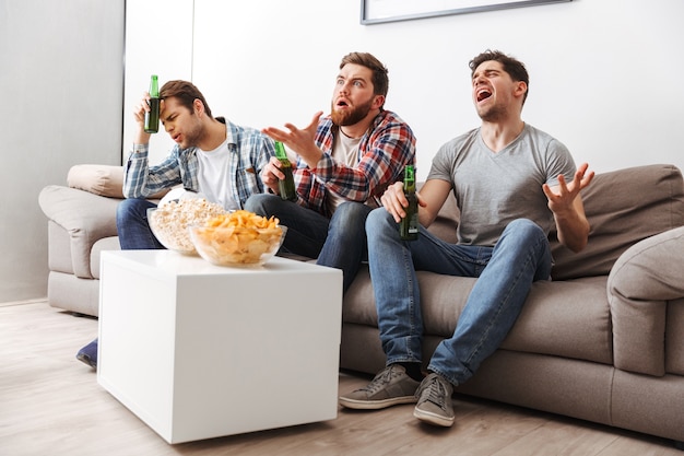 Portret van drie teleurgestelde jonge mannen die voetbal kijken terwijl ze thuis zitten met bier en snacks binnenshuis