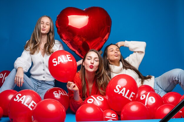 Portret van drie vrij lachende vriendinnen in casual kleding zitten met rode lucht ballonnen met verkoop- en procentteken