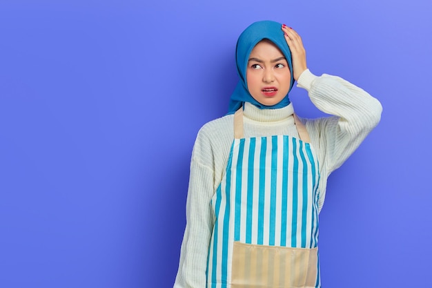 Portret van duizelige jonge huisvrouw vrouw in hijab en gestreepte schort opzij kijken terwijl handen op het hoofd geïsoleerd op paarse achtergrond mensen huisvrouw moslim levensstijl concept