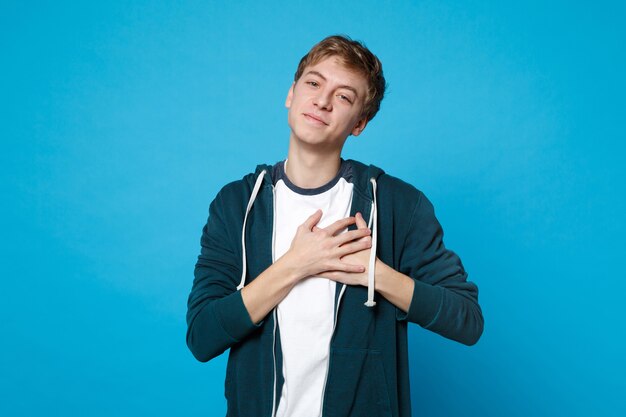 Portret van een aangename lachende jongeman in casual kleding met handpalmen gekruist op het hart, borst geïsoleerd op blauw.