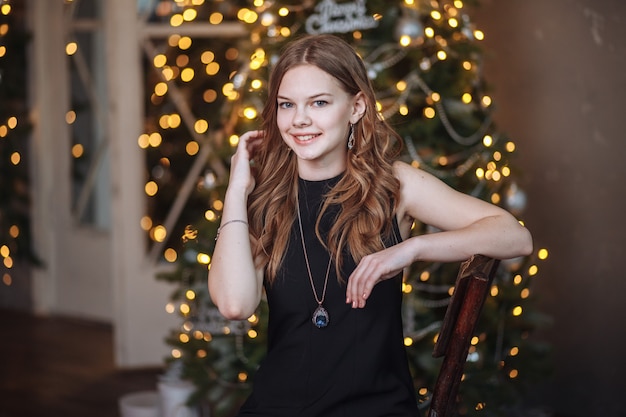 Portret van een aantrekkelijk jong meisje in een zwarte avondjurk tegen de achtergrond van een aangeklede kerstboom