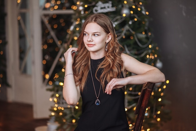 Portret van een aantrekkelijk jong meisje in een zwarte avondjurk tegen de achtergrond van een aangeklede kerstboom