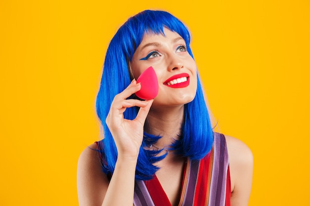 Portret van een aantrekkelijk lachend funky jong meisje met blauw haar met een casual outfit die geïsoleerd over een gele muur staat, make-upspons toont en wegkijkt