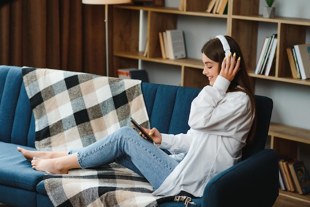 Portret van een aantrekkelijk meisje van 20 met een koptelefoon die een smartphone gebruikt en naar muziek luistert terwijl ze thuis op de bank zit