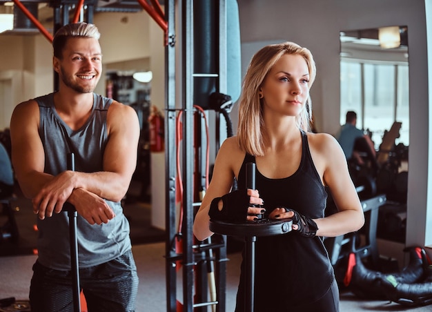 Portret van een aantrekkelijk sportief stel poseren terwijl ze op halters leunen, wegkijkend in de fitnessclub of sportschool.