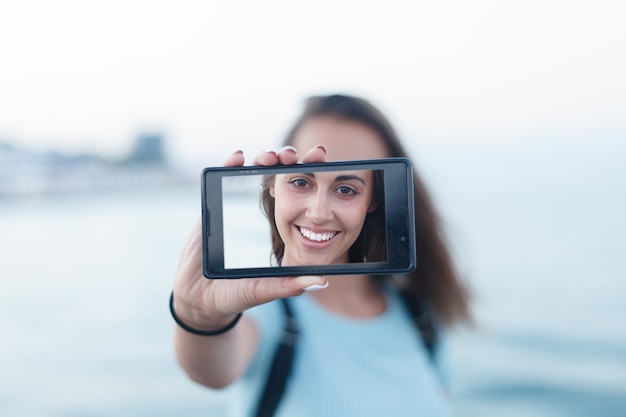 Portret van een aantrekkelijk tienermeisje dat op een zomerzandstrand op vakantie staat, met een smartphone-apparaat dat selfies van zichzelf maakt op vakantie tegen de blauwe hemel. Mensen reizen technologie.