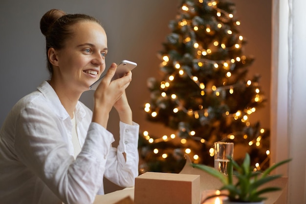 Portret van een aantrekkelijk vrolijk meisje dat december thuis doorbrengt met praten aan de telefoon, gefeliciteerd met haar vrienden die spraakberichten opnemen in een versierd huis binnenshuis