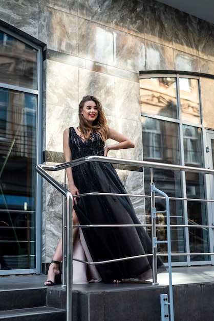 Portret van een aantrekkelijk vrouwelijk model in een prachtige zwarte avondjurk die zich voordeed in de straat van de stad, zomerdag