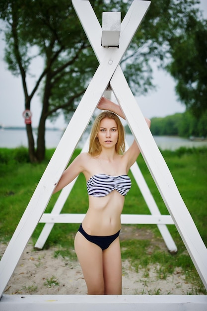 Foto portret van een aantrekkelijk vrouwelijk model poserend naast de driehoekige witte houten constructie in het park met een meer op de achtergrond