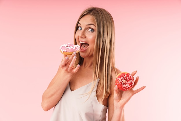 Foto portret van een aantrekkelijke blonde vrouw die een jurk draagt die lacht terwijl ze twee zoete donuts eet, geïsoleerd over een roze muur in de studio