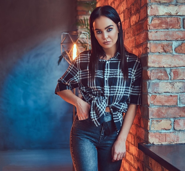 Portret van een aantrekkelijke brunette meisje gekleed in een flanellen shirt en jeans leunend op de muur in een kamer met loft interieur.