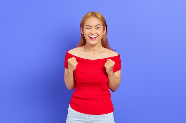 Portret van een aantrekkelijke en vrolijke jonge Aziatische vrouw met blond haar in een rode jurk die succes viert geïsoleerd op een paarse achtergrond