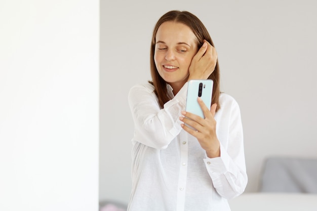 Portret van een aantrekkelijke gelukkige vrouw die een wit overhemd in casual stijl draagt, een mobiele telefoon in handen houdt, een selfie neemt, naar een smartphone kijkt en in een lichte kamer thuis poseert.