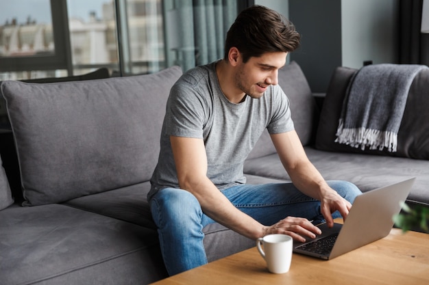 Portret van een aantrekkelijke glimlachende jonge, bebaarde man met vrijetijdskleding zittend op een bank in de woonkamer, met behulp van een laptopcomputer