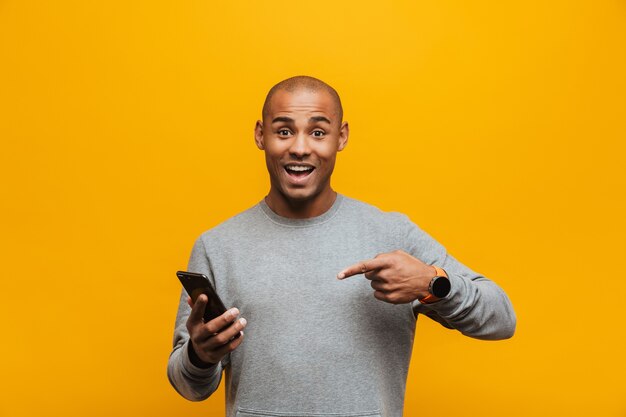 Portret van een aantrekkelijke glimlachende, zelfverzekerde, casual jonge afrikaanse man die over de gele muur staat en met de vinger naar de mobiele telefoon wijst