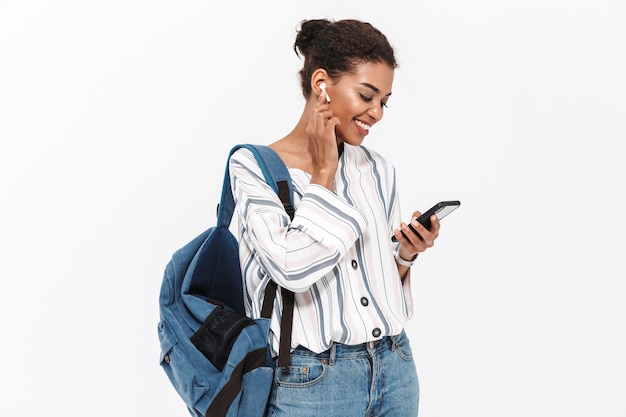 Portret van een aantrekkelijke jonge Afrikaanse vrouw met een rugzak die geïsoleerd over een witte muur staat, naar muziek luistert met draadloze oortelefoons, mobiele telefoon vasthoudt