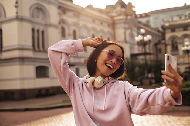 Portret van een aantrekkelijke jonge vrouw in een stijlvolle roze hoodie-koptelefoon en zonnebril glimlacht en neemt een selfie naar buiten