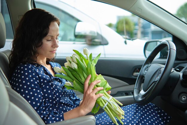 Portret van een aantrekkelijke jonge vrouw in een vrijetijdskleding die een boeket bloemen vasthoudt terwijl ze achter het stuur zit en een auto bestuurt