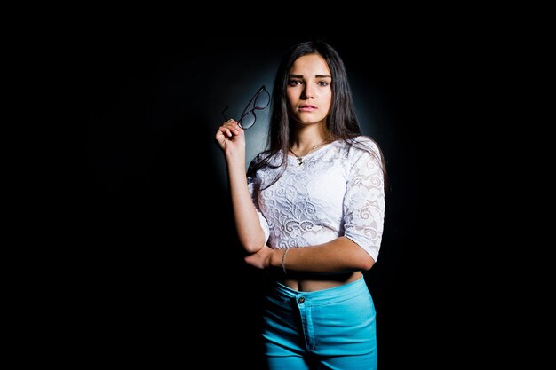 Portret van een aantrekkelijke jonge vrouw in witte top en blauwe broek poserend met haar bril in het donker