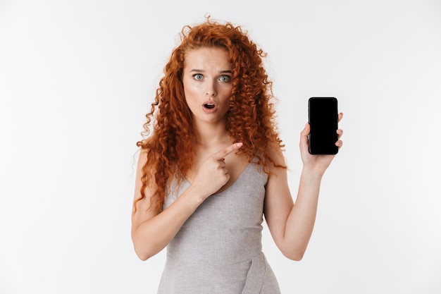 Portret van een aantrekkelijke jonge vrouw met lang krullend rood haar dat geïsoleerd staat en een lege mobiele telefoon toont