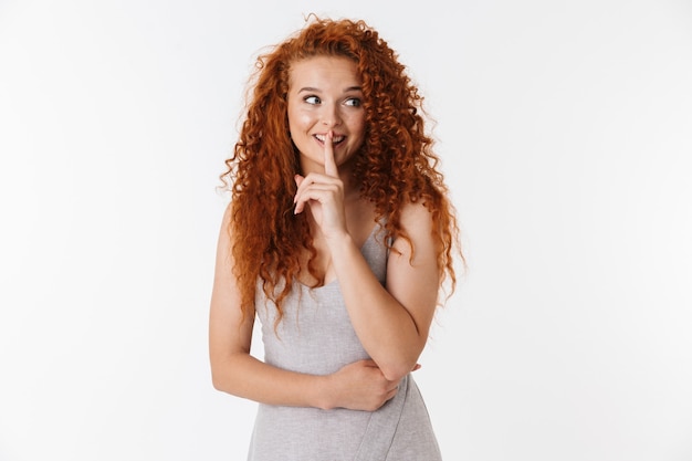 Portret van een aantrekkelijke jonge vrouw met lang krullend rood haar dat geïsoleerd staat en een stiltegebaar toont