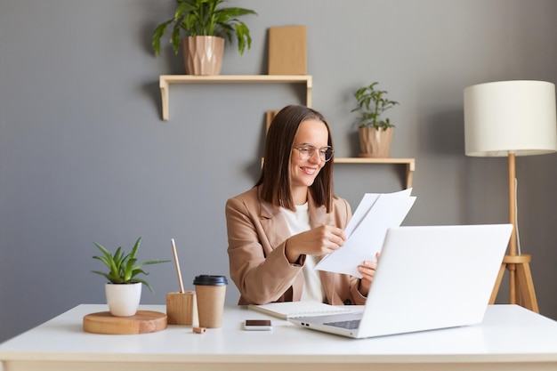 Portret van een aantrekkelijke lachende tevreden vrouw met bruin haar in een beige jas die online werkt op een laptop met papieren documenten in handen en geniet van haar werk