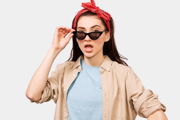 Portret van een aantrekkelijke, ondervraagde jonge vrouw die haar zwarte zonnebril afdoet en naar de camera kijkt met open mond, gekleed in een trendy outfit die zich voordeed op een witte achtergrond