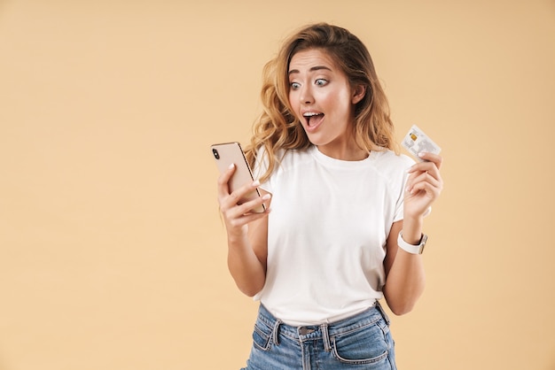 Portret van een aantrekkelijke opgewonden jonge vrouw met lang krullend blond haar met casual kleding die geïsoleerd staat, kijkend naar mobiele telefoon terwijl ze een plastic creditcard toont