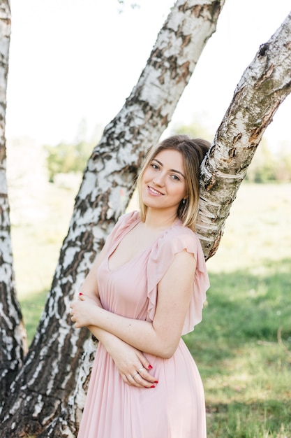 Portret van een aantrekkelijke romantische vrouw die een roze jurk draagt in de buurt van een berkenboom in het zomergroene park. vrouwelijk portret op lentelandschap.