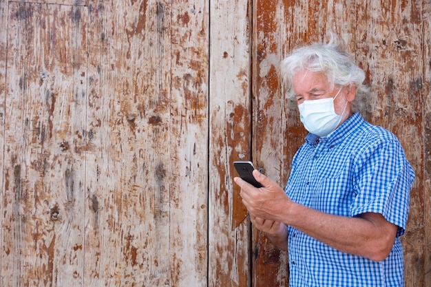 Portret van een aantrekkelijke senior man met wit haar die een gezichtsmasker draagt als gevolg van het coronavirus met behulp van een smartphone die tegen een houten deur staat - concept van gepensioneerde oudere technologie en sociaal