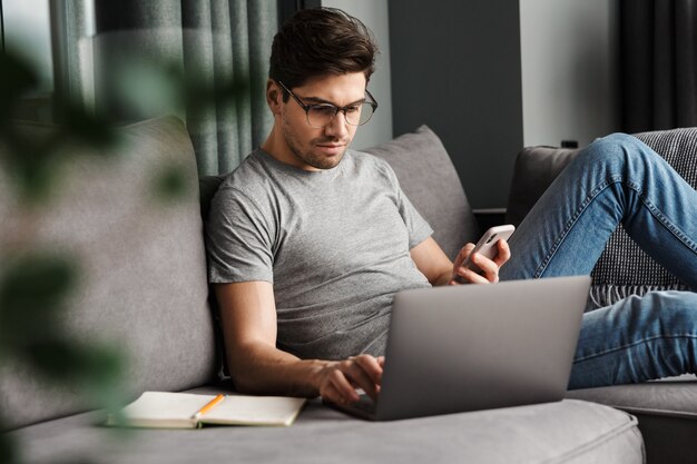 Portret van een aantrekkelijke serieuze jonge bebaarde man met vrijetijdskleding zittend op een bank in de woonkamer, met behulp van laptopcomputer