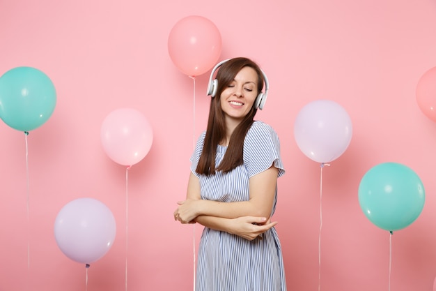 Portret van een aantrekkelijke tedere jonge vrouw met een koptelefoon in een blauwe jurk die muziek luistert en de handen gevouwen houdt op een pastelroze achtergrond met kleurrijke luchtballonnen. Verjaardagsfeestje concept.