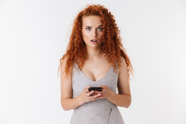Portret van een aantrekkelijke verwarde jonge vrouw met lang krullend rood haar die geïsoleerd staat, met behulp van mobiele telefoon