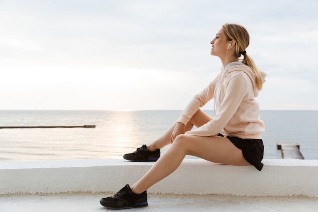 Portret van een aantrekkelijke vrouw die een trainingspak draagt en naar muziek luistert met een koptelefoon terwijl ze 's ochtends in de buurt van de kust zit