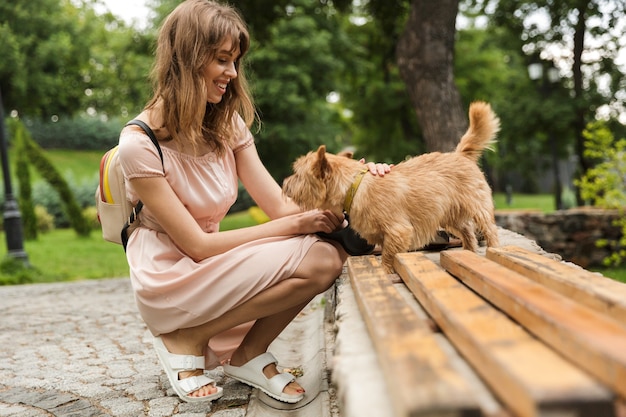 Portret van een aardige, tevreden vrouw met een rugzak die een hond aait terwijl ze in een park zit te hurken