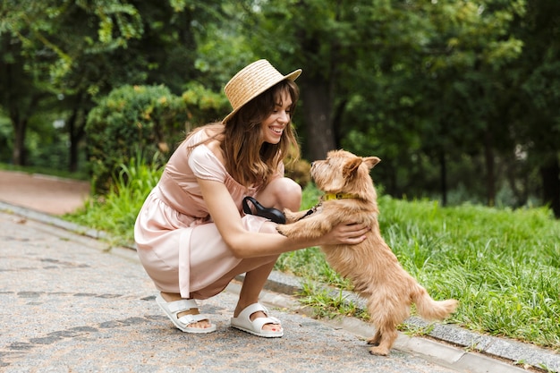 Portret van een aardige vrouw die een jurk draagt die een hond aait en glimlacht terwijl ze in een park zit te hurken