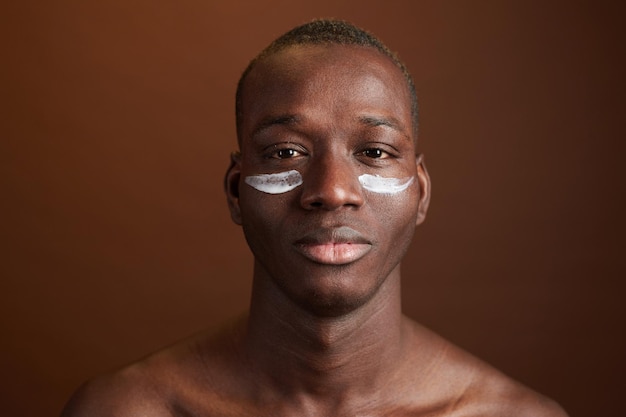 Portret van een Afrikaanse jongeman die de crème op zijn gezicht aanbrengt om de huid te beschermen die op een bruine achtergrond is geïsoleerd