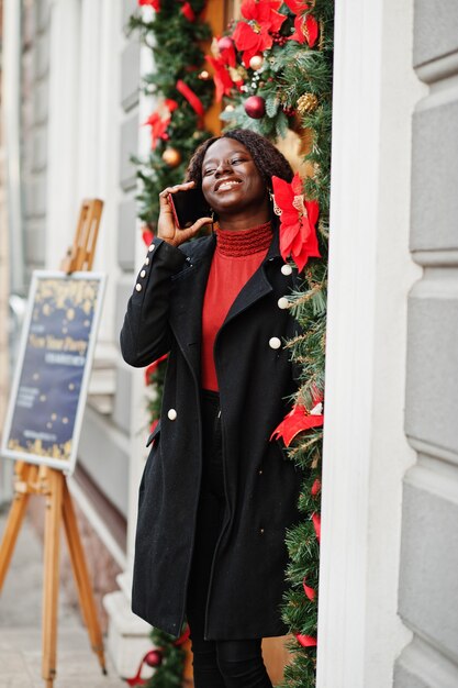 Portret van een Afrikaanse vrouw met krullend haar, gekleed in een modieuze zwarte jas en rode coltrui die buiten bij de deur staat met kerstversiering, oudejaarsavond. Spreek telefonisch.