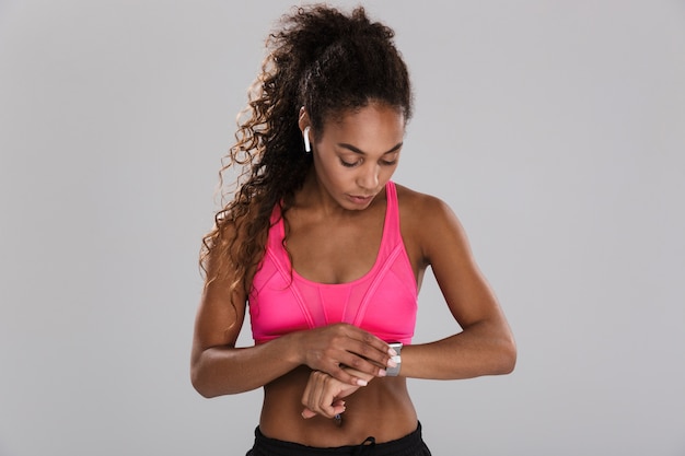 Portret van een afro-amerikaanse jonge sportvrouw geïsoleerd over grijze achtergrond, luisteren naar muziek met koptelefoon, kijken naar haar slimme horloge