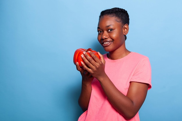 Portret van een Afro-Amerikaanse jonge vrouw met paprika die naar de camera kijkt die in de studio staat met een blauwe achtergrond. Tiener met gezonde voeding, groenten eten. Biologisch vegetarisch concept