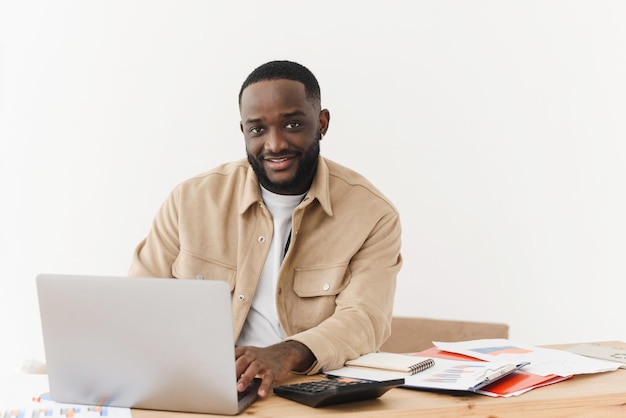 Portret van een Afro-Amerikaanse man die naar de camera kijkt terwijl hij aan tafel zit en op een laptop werkt